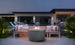 An outdoor seating area includes the sleek Halo Firepit by Ledge Lounger, surrounded by a beige sofa and cushions. Overlooking a pool and modern house with large windows under the twilight sky, green plants enhance the ambiance.