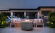An outdoor seating area includes the sleek Halo Firepit by Ledge Lounger, surrounded by a beige sofa and cushions. Overlooking a pool and modern house with large windows under the twilight sky, green plants enhance the ambiance.