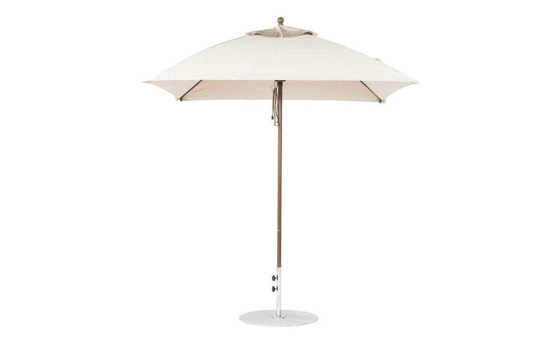 The Essential Fiberglass Umbrella by Ledge Lounger is a 7.5 square beige outdoor umbrella with a durable wooden pole and marine-grade fabrics, resting on a circular metal base, shown against a plain white background to emphasize style and durability.
