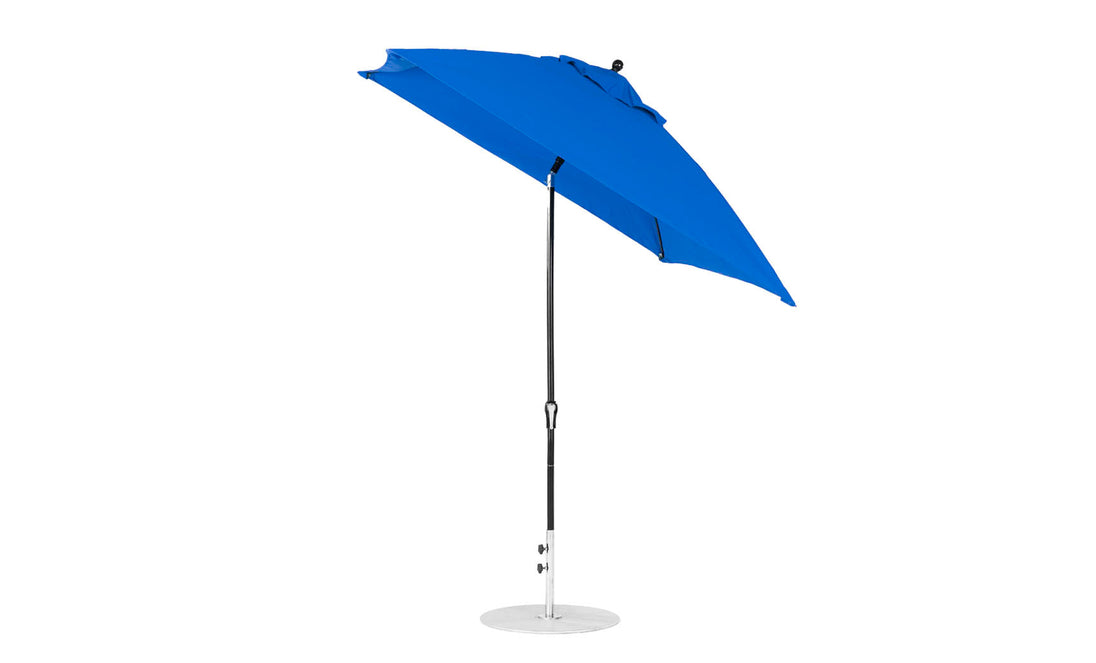 The Ledge Lounger Essential Fiberglass Umbrella features a blue, corrosion-resistant canopy with a black pole and round silver base. Partially open and tilted, its marine-grade fabric design includes a crank handle for easy adjustment against a plain white backdrop.