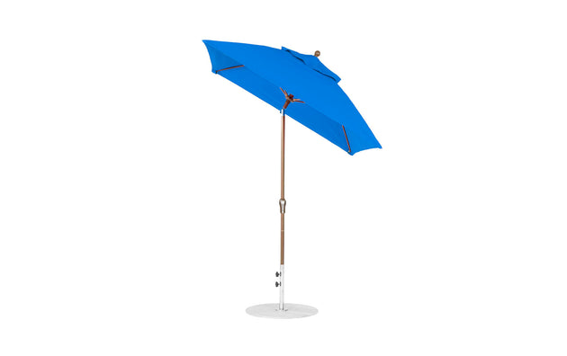 The Essential Fiberglass Umbrella by Ledge Lounger features a 6.5 square canopy made of marine-grade fabrics, highlighting a simple yet modern design. With its wooden pole and silver base, the open blue umbrella stands proudly upright.