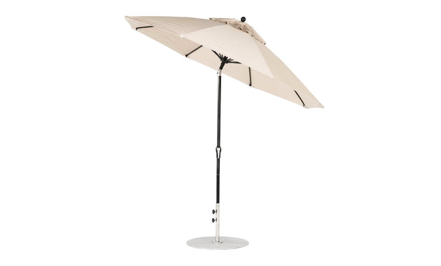 The open 9 beige Ledge Lounger Essential Fiberglass Umbrella, boasting marine-grade fabric and a central black pole, stands on a round metal base with its ribs and canopy slightly tilted against a white backdrop.
