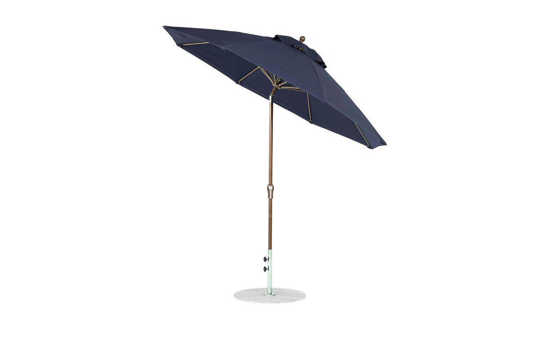 A 9 octagon Essential Fiberglass Umbrella by Ledge Lounger, featuring marine-grade fabrics, dark blue canopy with wooden pole on a white base, is open and tilted slightly to showcase its design.