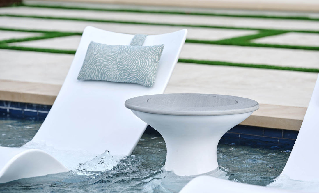 A modern poolside scene features two Ledge Lounger Autograph Headrest Pillows on white lounge chairs partially submerged in water. A round, white side table stands between them, set against a backdrop of landscaped grass and stone pathways.