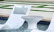 A modern poolside scene features two Ledge Lounger Autograph Headrest Pillows on white lounge chairs partially submerged in water. A round, white side table stands between them, set against a backdrop of landscaped grass and stone pathways.