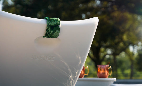 Close-up of a white Ledge Lounger chair with the Autograph Headrest Pillow, featuring a green towel on the backrest in an outdoor setting. Two copper mugs adorn a nearby table amidst sunlit greenery.