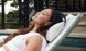 A woman with long brown hair relaxes on a poolside lounge chair, eyes closed, wearing a sleeveless white top. Her head is supported by an Autograph Headrest Pillow from Ledge Lounger. Behind her, a black metal fence and greenery complete the scene.