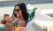 A person with long dark hair and sunglasses enjoys poolside relaxation in a green swimsuit with an Autograph Headrest Pillow by Ledge Lounger behind their head, smiling while holding a copper mug. Another individual with a similar mug is partially visible.