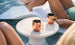 Two copper mugs with straws rest on a Ledge Lounger Autograph Standard Side Table. The poolside is lively, with people in green swimwear lounging by the water, exuding a relaxed and summery vibe.