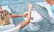 A person relaxes in the pool, placing a drink inside a cone-shaped Autograph Standard Side Table by Ledge Lounger. With its elegant design and secure lid, it serves as sophisticated floating storage. The clear water reflects the tables sleek look, while a white lounge chair is partially visible.