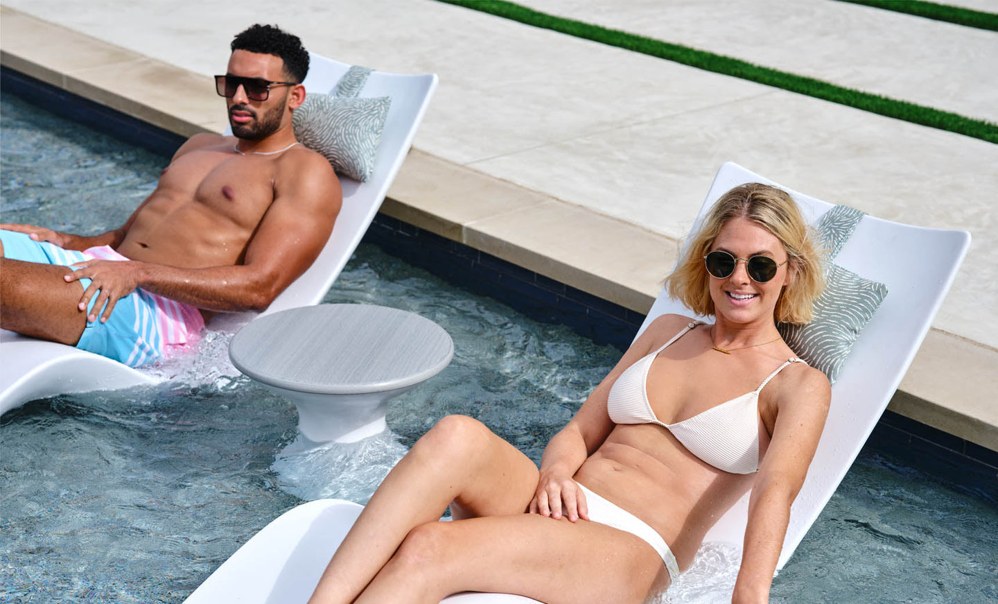 A man in colorful shorts and a woman in a white bikini, both wearing sunglasses, relax on white pool loungers partially submerged in water. Beside them is the sleek Autograph Standard Side Table by Ledge Lounger, complementing their sunny escape.