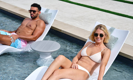 A man in colorful shorts and a woman in a white bikini, both wearing sunglasses, relax on white pool loungers partially submerged in water. Beside them is the sleek Autograph Standard Side Table by Ledge Lounger, complementing their sunny escape.