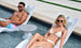 A man in colorful shorts and a woman in a white bikini, both wearing sunglasses, relax on white pool loungers partially submerged in water. Beside them is the sleek Autograph Standard Side Table by Ledge Lounger, complementing their sunny escape.