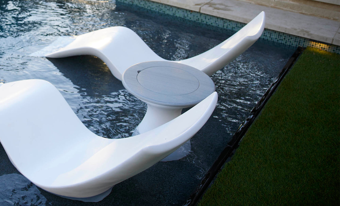 A sleek, white sculptural lounger set partially submerged in a shallow pool surrounds an Autograph Tall Ice Bin Side Table by Ledge Lounger. The pool is next to a grassy area and bordered by stone tiles, creating the perfect spot for relaxation and style.