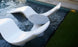 A sleek, white sculptural lounger set partially submerged in a shallow pool surrounds an Autograph Tall Ice Bin Side Table by Ledge Lounger. The pool is next to a grassy area and bordered by stone tiles, creating the perfect spot for relaxation and style.