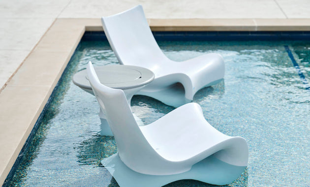 Two sleek Ledge Lounger white lounge chairs are partially submerged in a pool, flanking an Autograph Tall Ice Bin Side Table, creating a relaxing outdoor oasis.