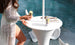 A woman in a lace robe holds a can by an outdoor table near the pool. On the table, next to a Ledge Lounger Autograph Umbrella Stand Ice Bin, sits another can and some magazines. The scene is bright and summery.