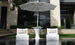 Two modern white lounge chairs in water face a Ledge Lounger Autograph Umbrella Stand Ice Bin and tall outdoor table. A drink and fruit are on the table, set outside a contemporary building with large windows and lush greenery.