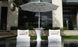 Two modern white lounge chairs in water face a Ledge Lounger Autograph Umbrella Stand Ice Bin and tall outdoor table. A drink and fruit are on the table, set outside a contemporary building with large windows and lush greenery.