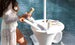 A person in a white lace cover-up places a can into the Ledge Lounger Autograph Umbrella Stand Ice Bin, a white outdoor table with storage that doubles as an ice bin. Another can and some magazines sit beside it, perfectly positioned by the pools clear blue water.