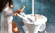 A person in a white lace cover-up places a can into the Ledge Lounger Autograph Umbrella Stand Ice Bin, a white outdoor table with storage that doubles as an ice bin. Another can and some magazines sit beside it, perfectly positioned by the pools clear blue water.