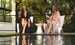 Two women sit in Classic Luxury In-Pool Chairs by Ledge Lounger, wearing dark and light floral robes. They hold wine glasses and laugh by the pool, surrounded by trees, creating a relaxed and cheerful outdoor scene.