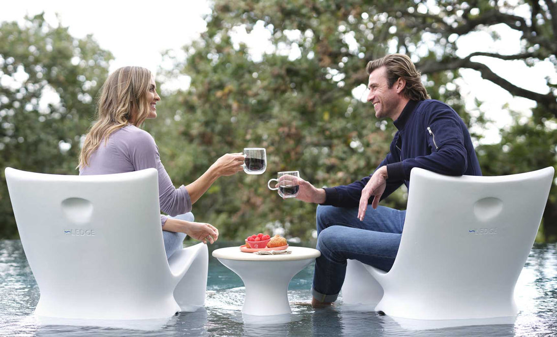A woman and man relax on Ledge Loungers Classic Luxury In-Pool Chair Duo, facing each other with drinks in hand. An Autograph side table holds their snacks, while trees create a scenic backdrop for their casual conversation in the pools shallow end.