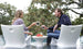 A woman and man relax on Ledge Loungers Classic Luxury In-Pool Chair Duo, facing each other with drinks in hand. An Autograph side table holds their snacks, while trees create a scenic backdrop for their casual conversation in the pools shallow end.