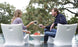 A woman and man relax on Ledge Loungers Classic Luxury In-Pool Chair Duo, facing each other with drinks in hand. An Autograph side table holds their snacks, while trees create a scenic backdrop for their casual conversation in the pools shallow end.