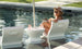 A person lounges in a Ledge Lounger Relaxation In-Pool Chair & Ice Bin Bundle - Lowback, partially submerged in clear waters. Wearing a hat and sunglasses, theyre reading a magazine with two glasses on a small table between the chairs, creating the perfect sunny and relaxed setting.