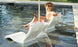 The woman in a white swimsuit and sun hat sits comfortably on a Ledge Lounger Relaxation In-Pool Chair & Ice Bin Bundle - Lowback, partially submerged, smiling with a magazine. Glasses rest on the nearby table in her sunny poolside oasis.