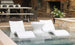 A pair of Ledge Loungers Outdoor Oasis In-Pool Furniture Bundles, featuring deep chaise loungers in white, are partially submerged in a shallow pool. An ice bin side table sits nearby against a stone wall with plants and a wooden dining table set in the background.