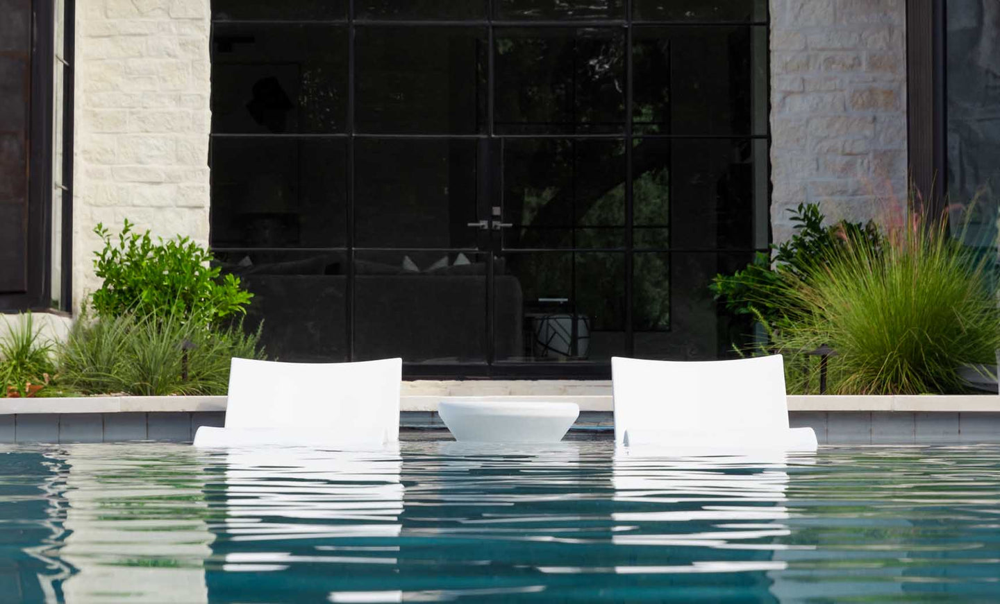 Two white Outdoor Oasis In-Pool Furniture Bundle loungers by Ledge Lounger sit partially submerged in the pool, facing a modern building with large glass windows. The clear water reflects the loungers, offering an exquisite escape amidst lush greenery.