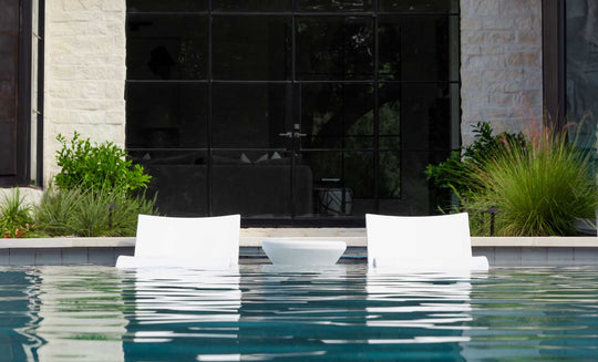 Two white Outdoor Oasis In-Pool Furniture Bundle loungers by Ledge Lounger sit partially submerged in the pool, facing a modern building with large glass windows. The clear water reflects the loungers, offering an exquisite escape amidst lush greenery.