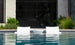 Two white Outdoor Oasis In-Pool Furniture Bundle loungers by Ledge Lounger sit partially submerged in the pool, facing a modern building with large glass windows. The clear water reflects the loungers, offering an exquisite escape amidst lush greenery.