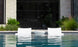 Two white Outdoor Oasis In-Pool Furniture Bundle loungers by Ledge Lounger sit partially submerged in the pool, facing a modern building with large glass windows. The clear water reflects the loungers, offering an exquisite escape amidst lush greenery.