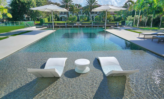 The Outdoor Oasis In-Pool Furniture Bundle by Ledge Lounger showcases luxurious in-pool chaise loungers with a round table, shaded by umbrellas amidst greenery and palm trees. A large house with a veranda completes the perfect poolside essentials setup.