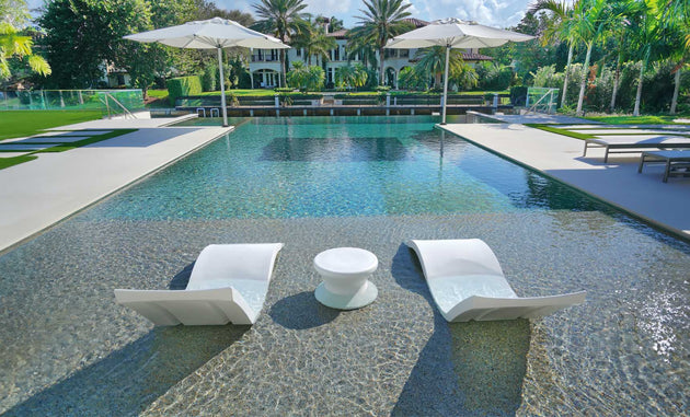 The Outdoor Oasis In-Pool Furniture Bundle by Ledge Lounger showcases luxurious in-pool chaise loungers with a round table, shaded by umbrellas amidst greenery and palm trees. A large house with a veranda completes the perfect poolside essentials setup.