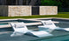 Two Ledge Lounger white in-pool chaise loungers with pillows, accompanied by a small round table, are semi-submerged in a shallow pool. A stone wall and grassy lawn add to the scenes tranquility. Enjoy the sunny escape with the Outdoor Paradise In-Pool Furniture Bundle.