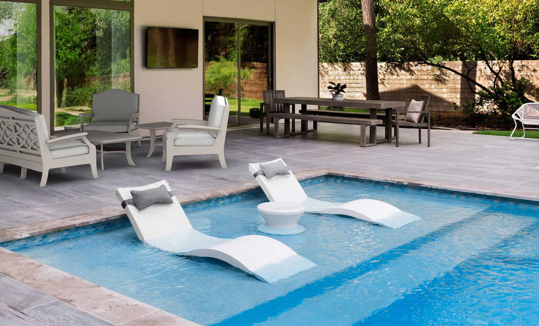 Experience an outdoor paradise with the Ledge Lounger Outdoor Paradise In-Pool Furniture Bundle: two Signature In-Pool Chaise Loungers elegantly flank a sleek round side table, set amid a cozy seating area and dining space on gray tiles, framed by lush trees.