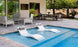Experience an outdoor paradise with the Ledge Lounger Outdoor Paradise In-Pool Furniture Bundle: two Signature In-Pool Chaise Loungers elegantly flank a sleek round side table, set amid a cozy seating area and dining space on gray tiles, framed by lush trees.