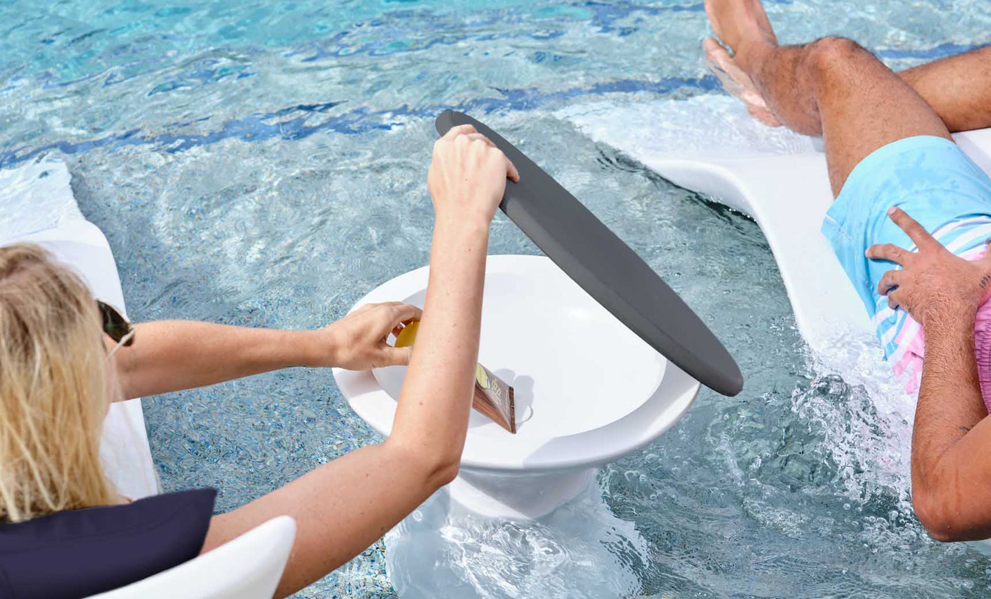 A person adjusts a black, round shade over their drink on a floating tray while enjoying the Resort Luxury Outdoor Paradise In-Pool Furniture Bundle by Ledge Lounger. Another relaxes on a chaise lounger in the water, embracing poolside leisure and sunshine.