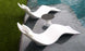 The Resort Luxury Outdoor Paradise In-Pool Furniture Bundle from Ledge Lounger features two wave-shaped, modern white chaise loungers partially submerged in a pool beside a small, black-topped round table, creating a serene and sunny poolside retreat where the pool meets the grassy lawn.