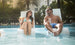 A man and woman unwind on Ledge Loungers Relaxation In-Pool Chair & Ice Bin Bundle. She savors a cocktail while he has a soda, both clad in sunglasses and swimwear, with palm trees and a building completing their idyllic poolside scene.