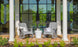 Two gray rocking chairs on a brick porch have a Ledge Lounger Affinity Side Table between them. Reflections of the garden greenery in a glass door enhance the scene, while a white column and potted plants frame this inviting outdoor area, highlighting chic accessory potential.