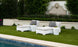 Two white lounge chairs with gray cushions and Ledge Lounger Affinity Side Tables are placed on a green lawn by the pool. A beige sun hat is on one chair. The setting includes a stone wall with a carved lions head and lush greenery, forming an inviting oasis for relaxation.