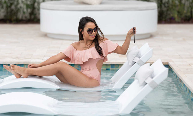 A woman in a pink off-shoulder swimsuit relaxes on a white pool float in the swimming pool, smiling and enjoying her drink with sunglasses on. In the background, the scene is enhanced by Ledge Loungers Outdoor Escape In-Pool Furniture Bundle, featuring outdoor elements like sunbeds and greenery.