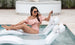 A woman in a pink off-shoulder swimsuit relaxes on a white pool float in the swimming pool, smiling and enjoying her drink with sunglasses on. In the background, the scene is enhanced by Ledge Loungers Outdoor Escape In-Pool Furniture Bundle, featuring outdoor elements like sunbeds and greenery.
