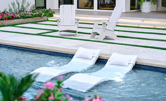 The Outdoor Escape In-Pool Furniture Bundle by Ledge Lounger features two white Signature Chaise lounge chairs with water jets for relaxation and two Adirondack chairs on a stone patio with grass accents, all bordered by pink flowers and greenery.