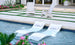 The Outdoor Escape In-Pool Furniture Bundle by Ledge Lounger features two white Signature Chaise lounge chairs with water jets for relaxation and two Adirondack chairs on a stone patio with grass accents, all bordered by pink flowers and greenery.